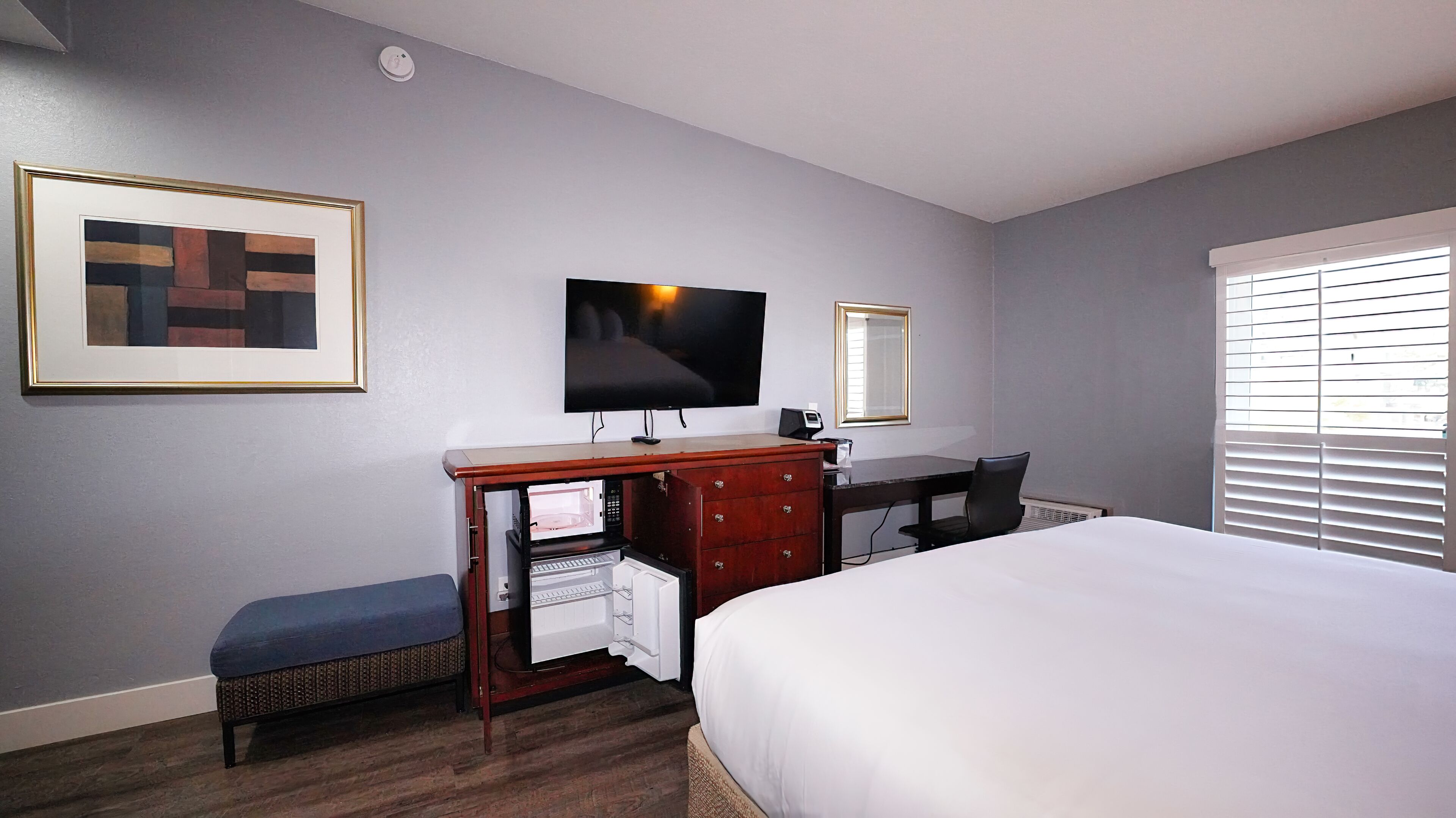 A hotel room with a bed, a desk with a computer, a television, a framed picture, and a window with blinds.