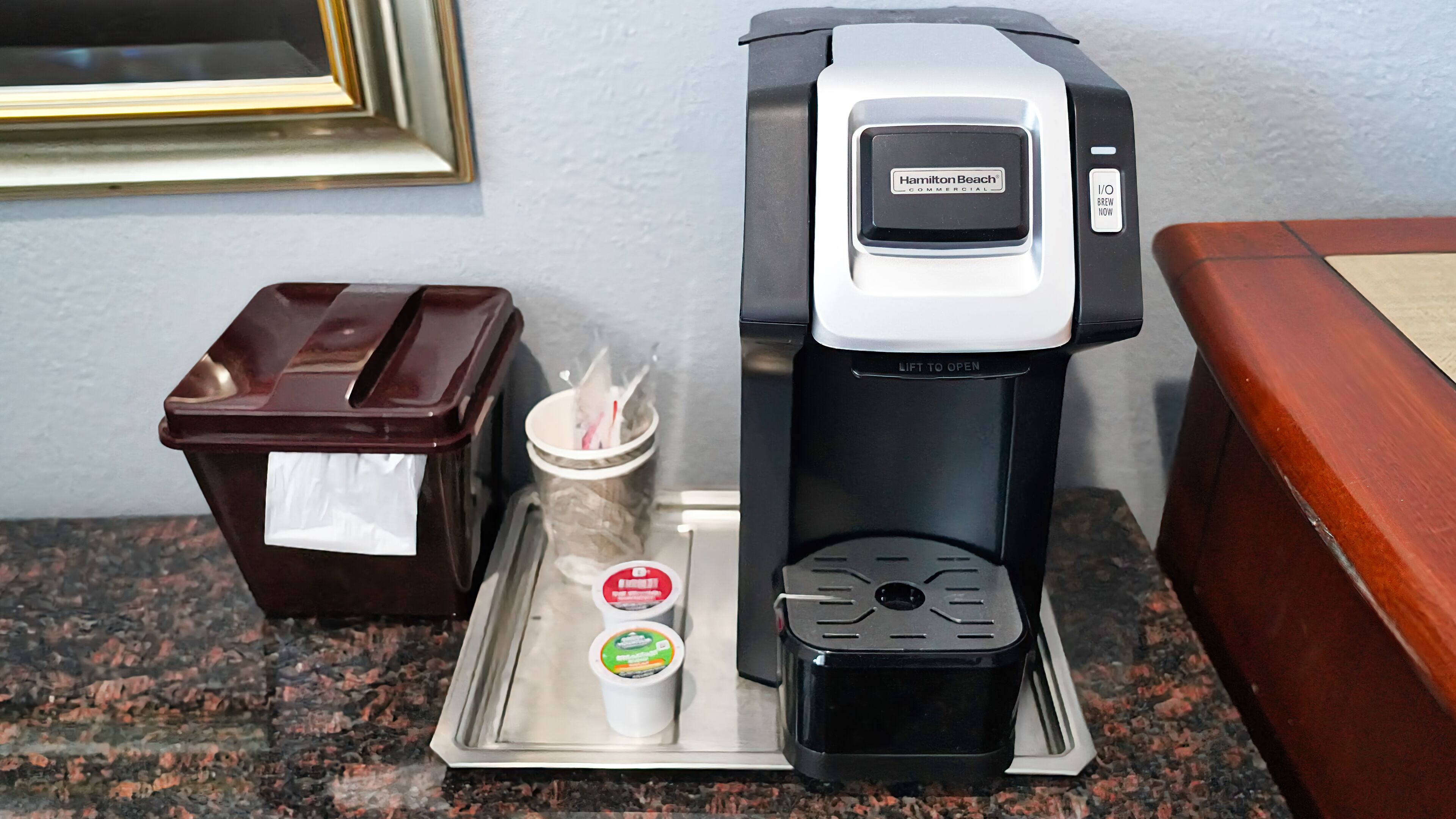 Uma cafeteira com a etiqueta Hamilton Beach, uma lixeira e um tabuleiro com duas chávenas de café e um frasco de açúcar.
