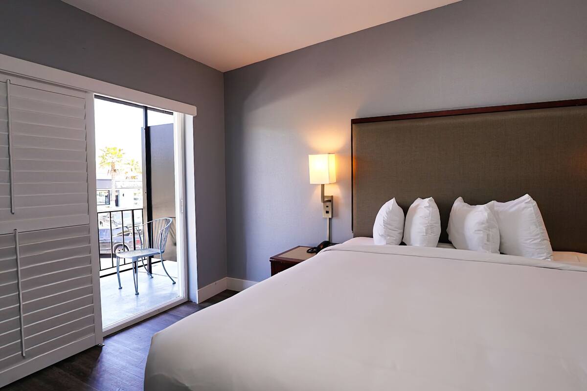 A hotel room with a large bed, two bedside lamps, a nightstand, and a sliding glass door leading to a balcony with a chair.