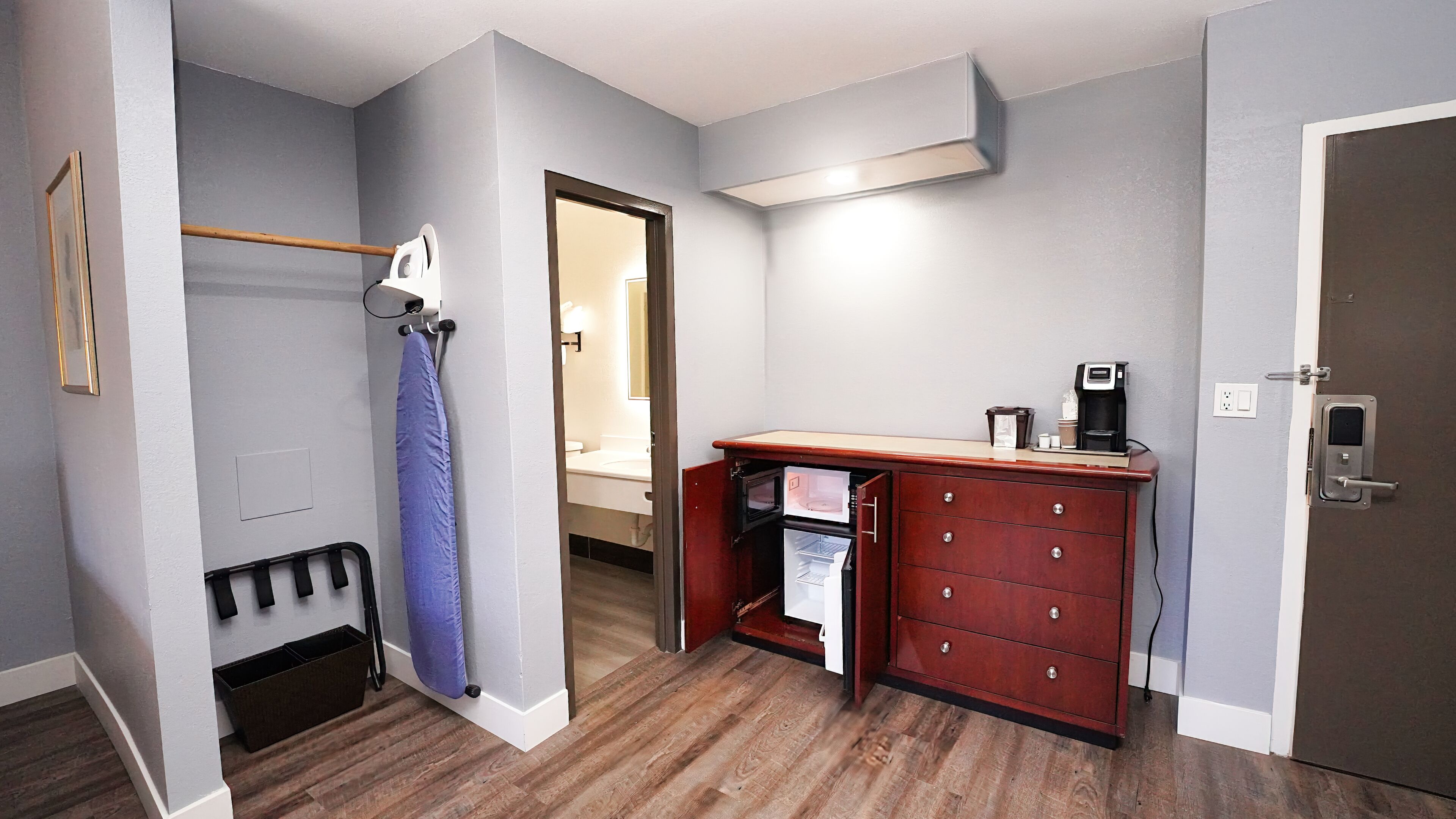 Habitación compacta con suelo de madera, escritorio integrado con cafetera, un refrigerador pequeño y zona de ducha.