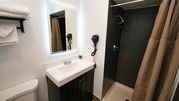 Un baño con lavamanos blanco, espejo, secador de pelo y ducha con pared de azulejos oscuros.