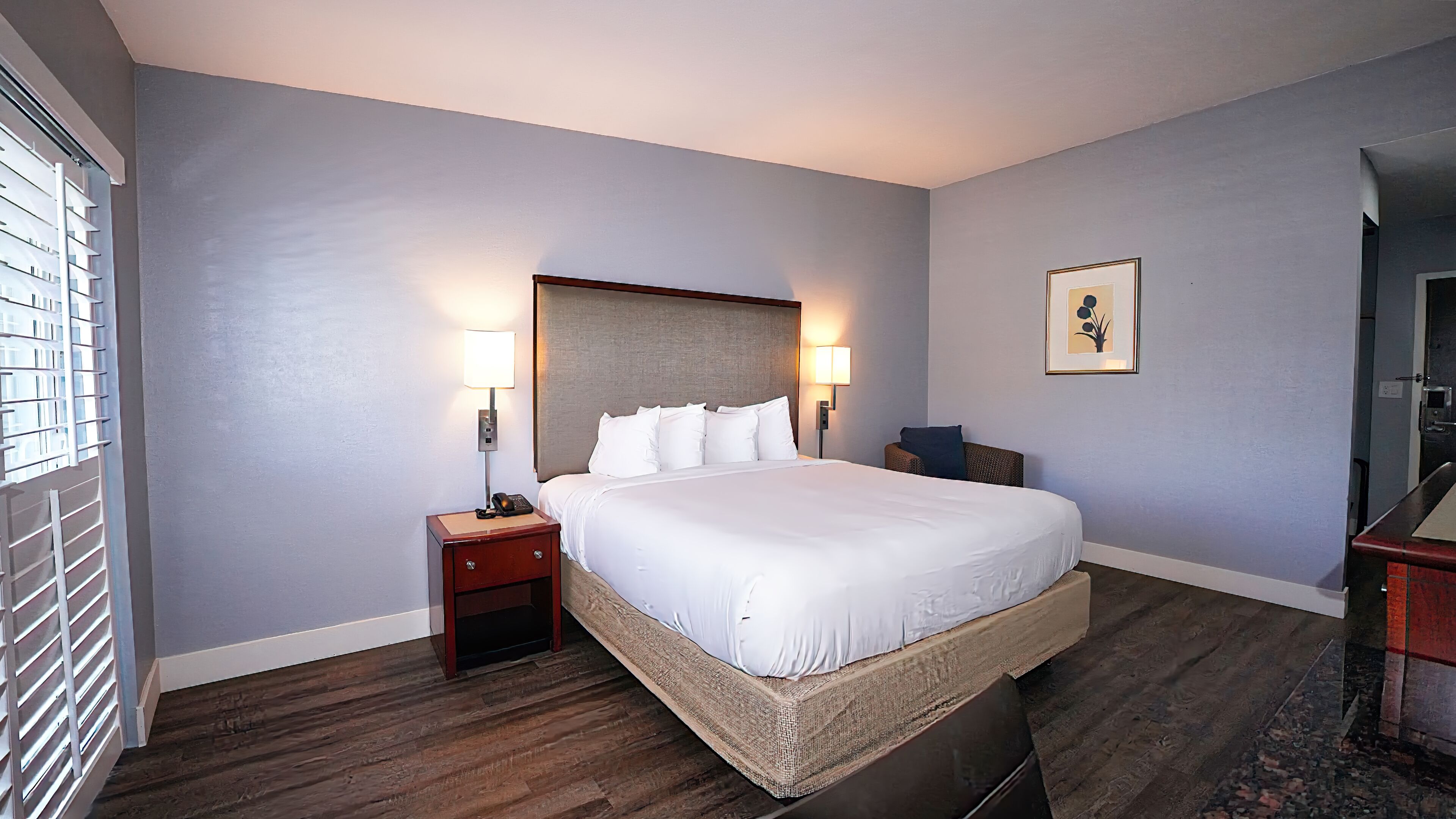Habitación de hotel con una cama grande, dos mesitas de noche con lámparas, un cuadro enmarcado en la pared y suelo de madera.