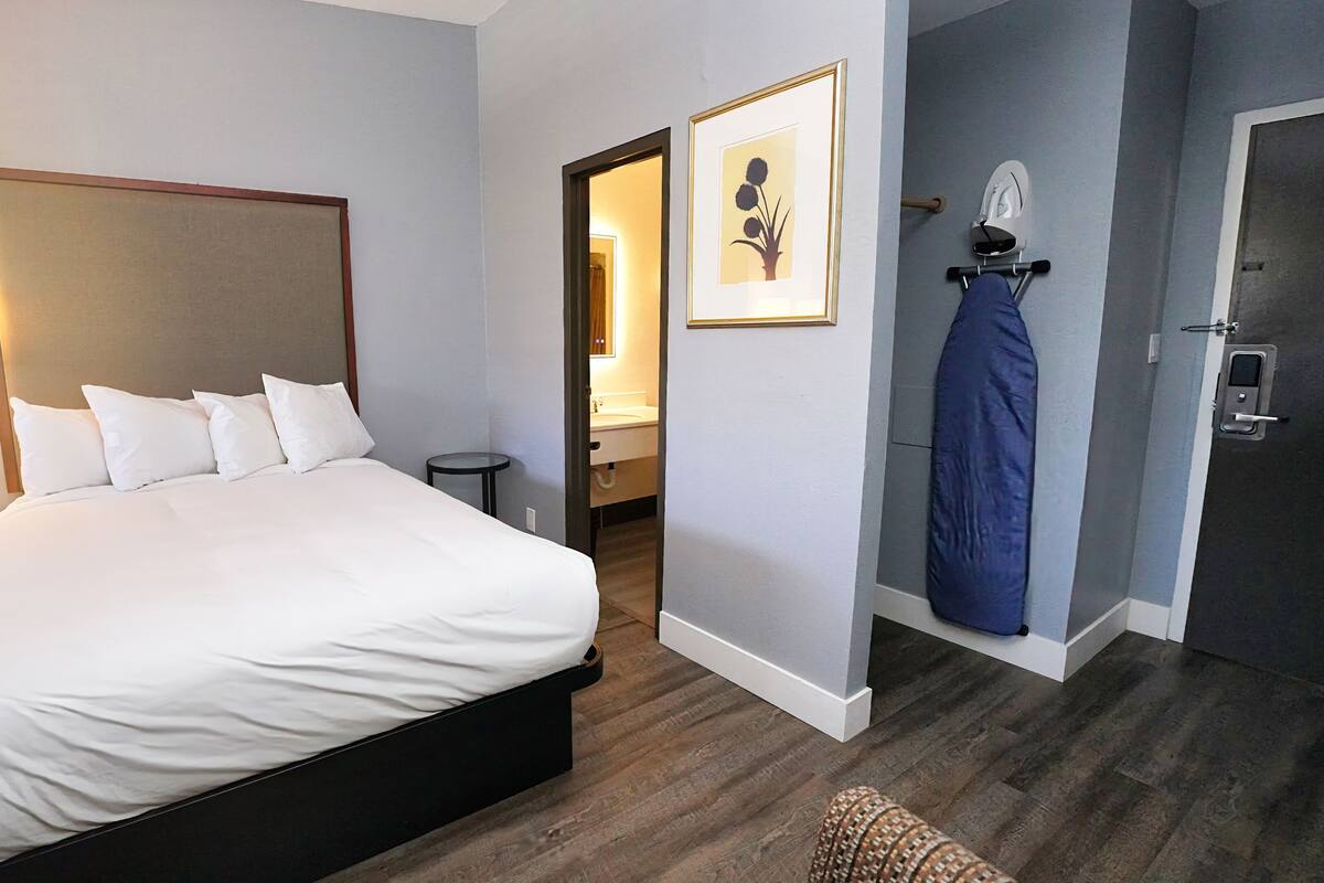 A hotel room with a large bed, bedside table with a phone, a small round table, a bathroom visible through an open door, and a coat rack with a jacket.