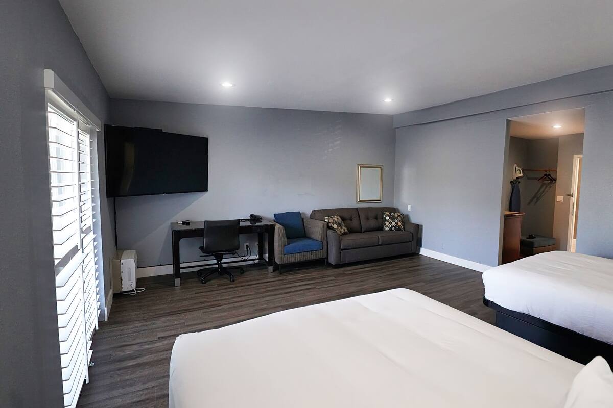 A hotel room with two beds, a desk, a sofa, a TV, and a shower area.