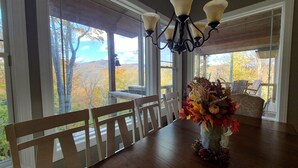 Un comedor con mesa de madera, sillas, una araña y vistas del follaje otoñal a través de amplios ventanales.