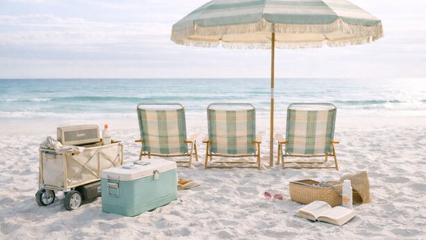 Een strandscène met een geribbelde strandparasol, twee strandstoelen, een koelbox, een koffer en een mand met zonnebril en een boek.