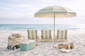 Een strandscène met een geribbelde strandparasol, twee strandstoelen, een koelbox, een koffer en een mand met zonnebril en een boek.