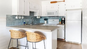 Een moderne keuken met witte kasten, een ingebouwde magnetron, een fornuis en een koelkast. Er staan twee rieten barkrukken en een houten spatel boven de aanrecht.