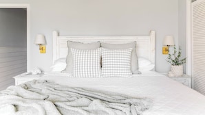 Een slaapkamer met een wit bed, geruit kussens, twee bedlampen, een witte nachtkastje en een witte muur.