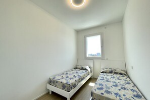 Une petite pièce lumineuse, avec deux lits simples, une fenêtre et un luminaire au plafond.