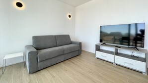 A modern living room with a grey sofa, a flat-screen TV on a white TV stand, and two recessed ceiling lights. - Appartamento Blu Homes 2 - Carraro Immobiliare Jesolo (Lido di Jesolo)