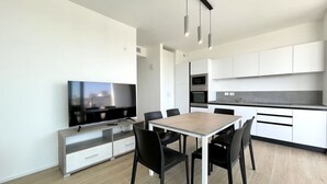 A modern kitchen with white cabinets, a dining area with a wooden table and black chairs, and a flat-screen TV on a white cabinet.