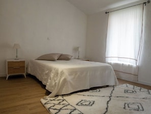 A bedroom with a bed, two bedside tables, a window with curtains, and a patterned rug. - Charming village house in the heart of the Luberon (Oppède)