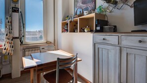 Una cocina con ventana, una mesa, sillas, un radiador, un reloj y una estantería con diversos objetos.