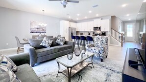 A modern living room with a grey sofa, a coffee table, and a patterned armchair. There is a kitchen area with white cabinets and a microwave. - Private Pool • FlowRider Surf Simulator • 3 Ensuites • 15 Min to Disney (Kissimmee)