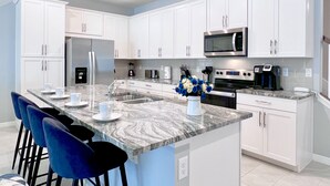 A modern kitchen with white cabinets, a marble countertop, and a built-in oven. There are bar stools and a vase of flowers on the counter. - Private Pool • FlowRider Surf Simulator • 3 Ensuites • 15 Min to Disney (Kissimmee)