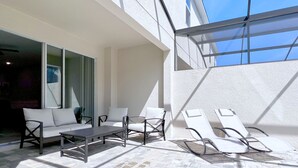 A sunlit outdoor patio with a sofa, chairs, and a table, featuring a view of the sky through a glass structure. - Private Pool • FlowRider Surf Simulator • 3 Ensuites • 15 Min to Disney (Kissimmee)