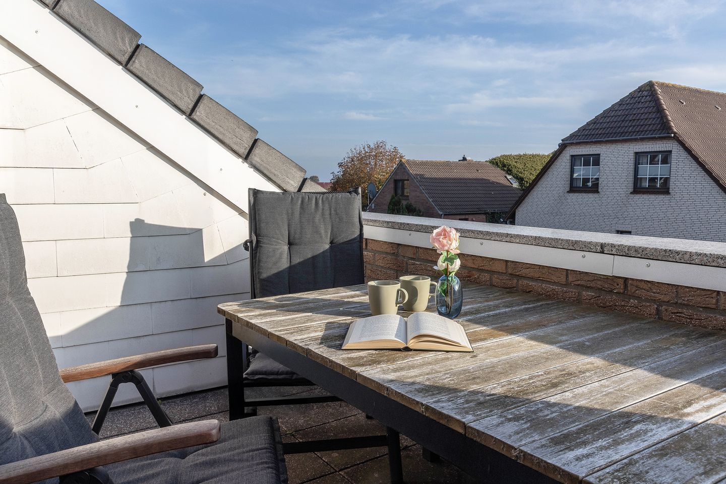 A wooden deck with a table, two chairs, a book, a vase with a flower, and two mugs.