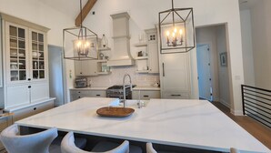 Une cuisine moderne avec des meubles blancs, un grand îlot central doté de tabourets, des luminaires suspendus et une cuisinière visible.