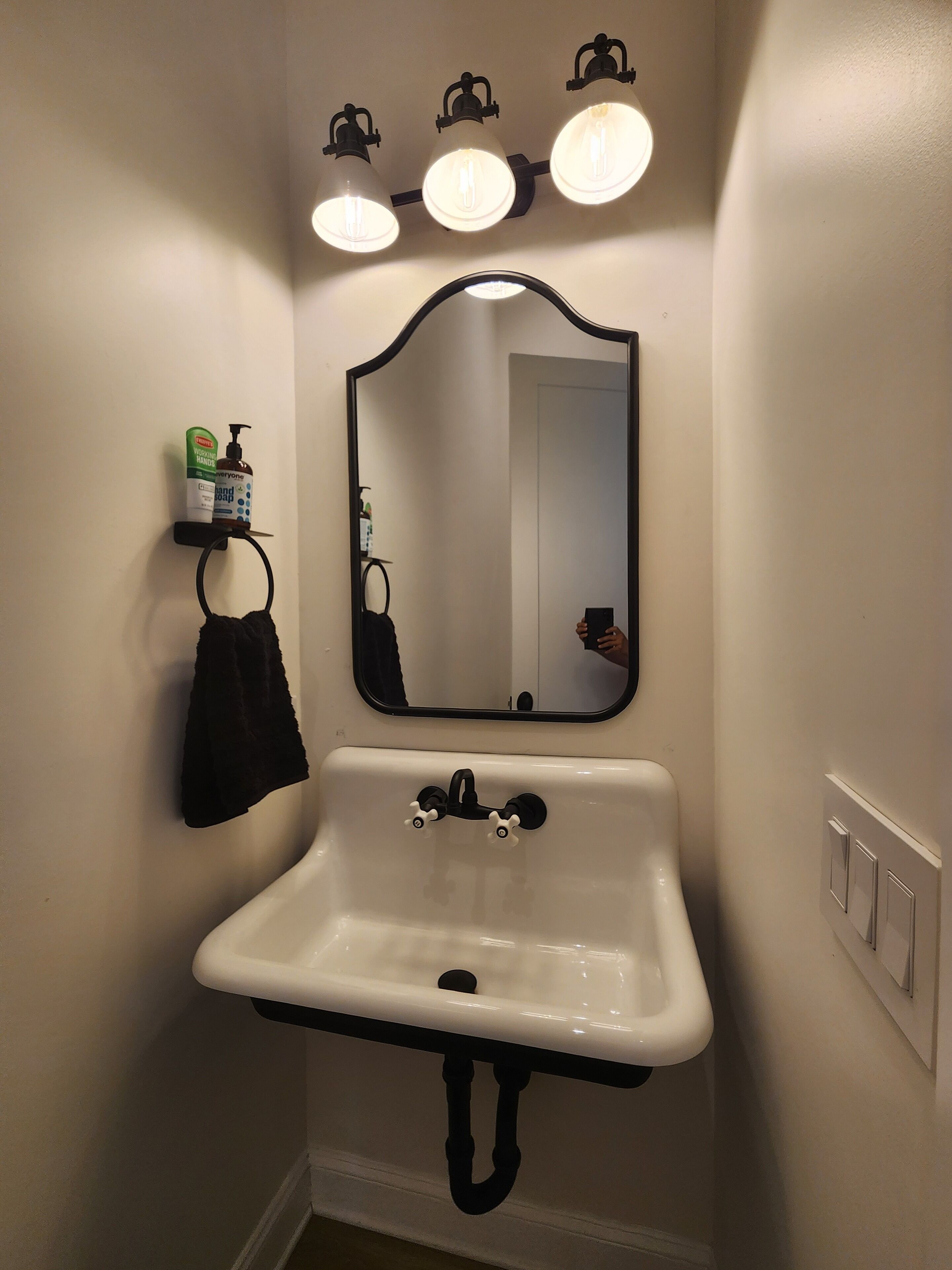 A bathroom with a white sink, three wall-mounted lights, a mirror, and a towel rack with a black towel.