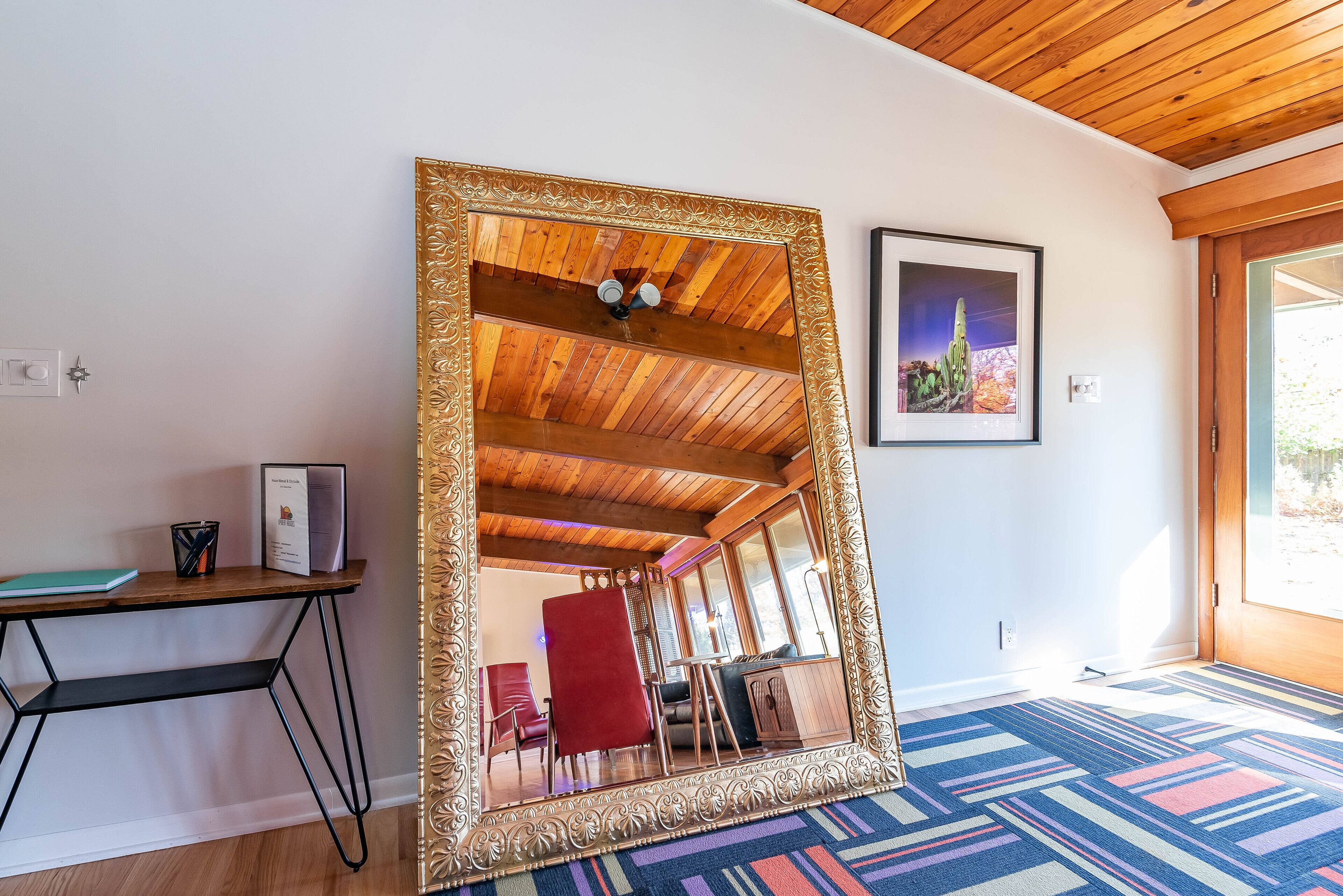 A room with a large mirror, wooden ceiling, a desk with a chair, a colorful rug, and a framed picture on the wall.