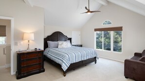 Une chambre avec un grand lit, une commode, un ventilateur de plafond et une fenêtre donnant sur les arbres.