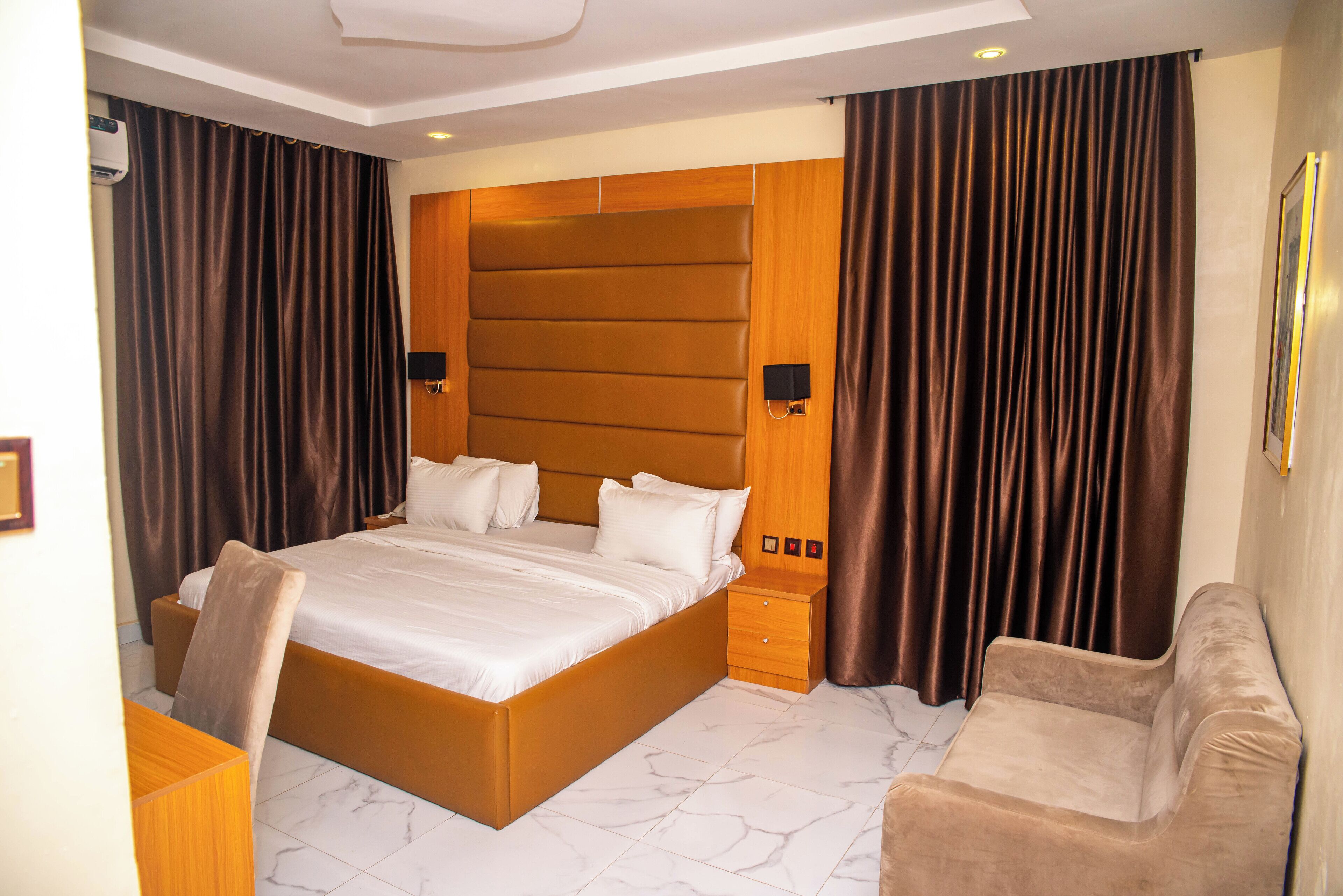 A modern hotel room with a large bed, wooden headboard, two bedside tables, a desk, and a comfortable armchair.