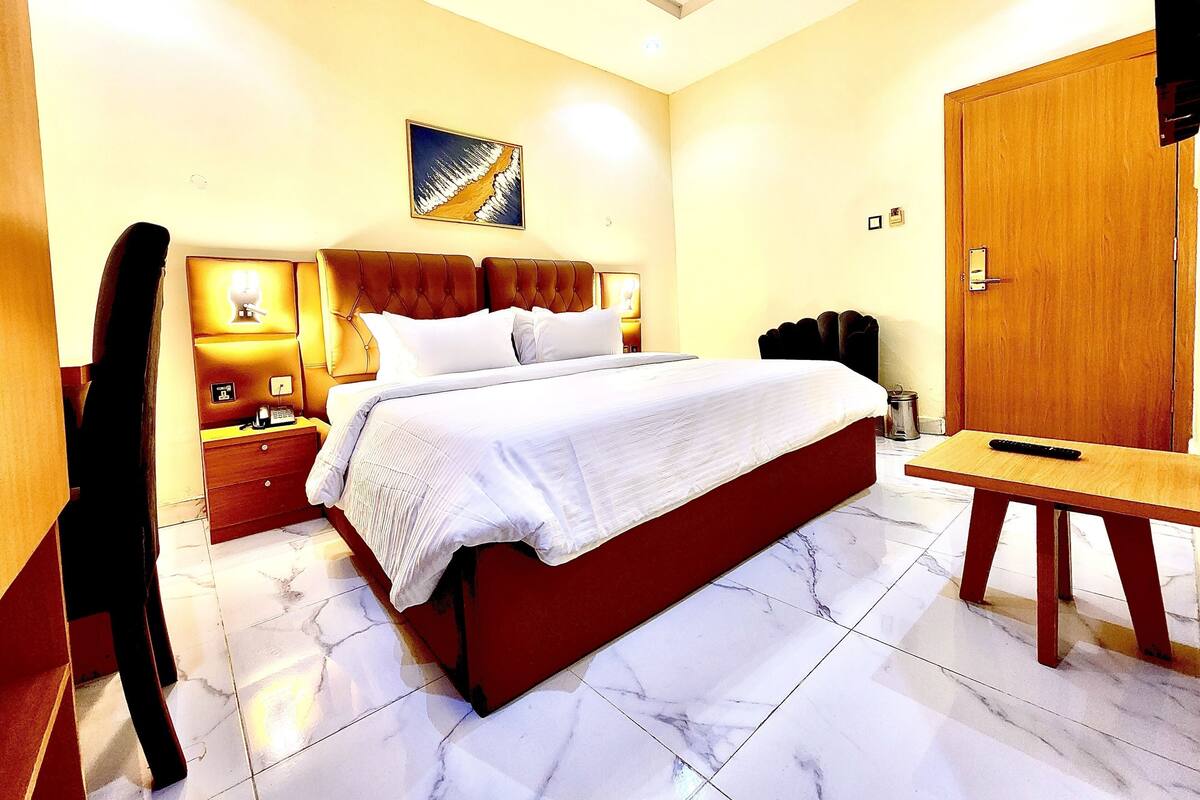 A hotel room with a bed, bedside table, lamp, and a painting on the wall.