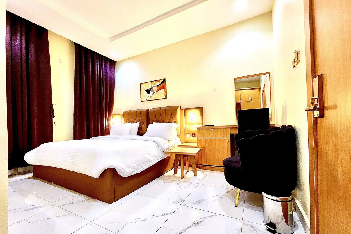 A hotel room with a large bed, a desk, a chair, a wardrobe, and a mirror.