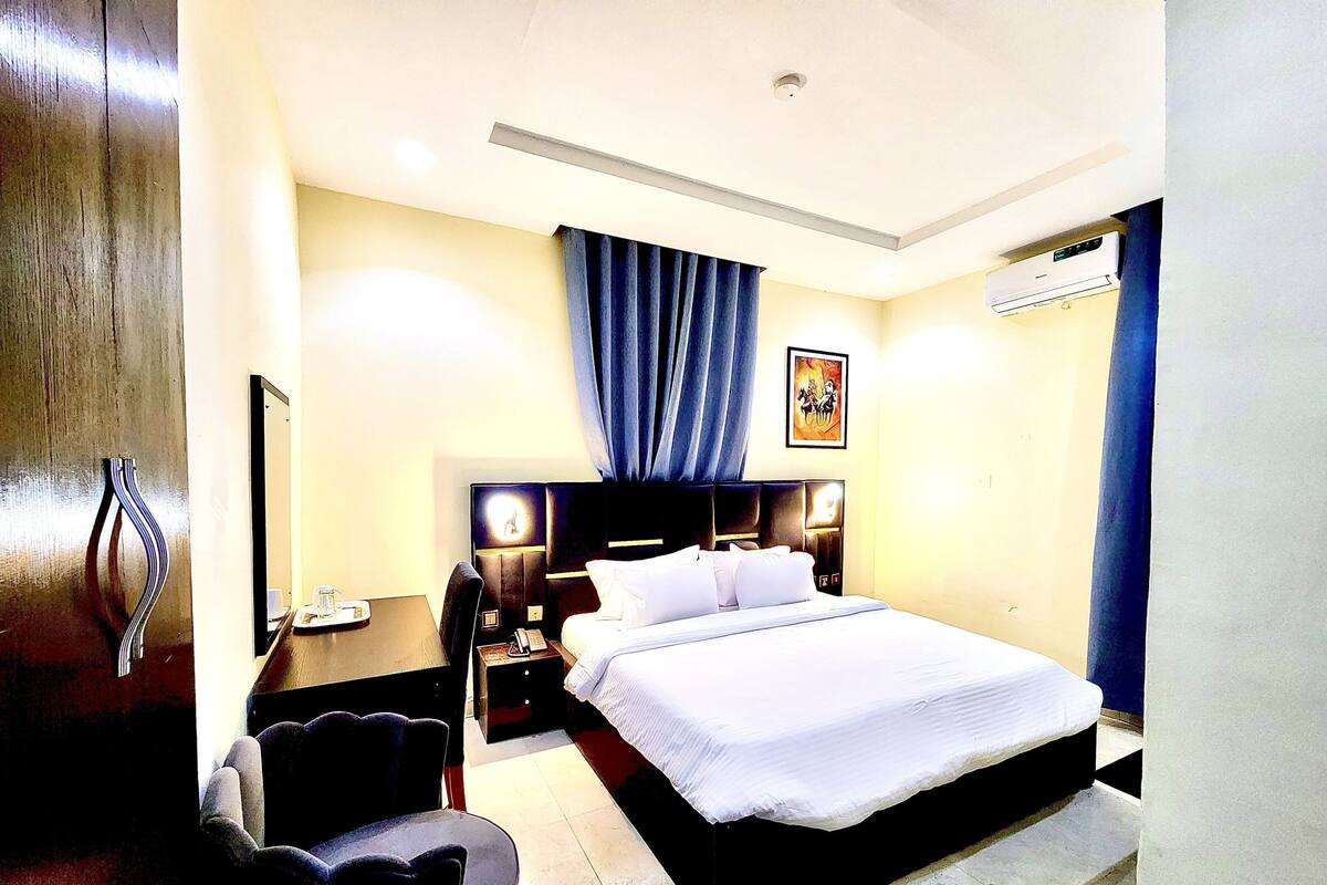 A hotel room with a bed, bedside table, chair, and a painting on the wall.