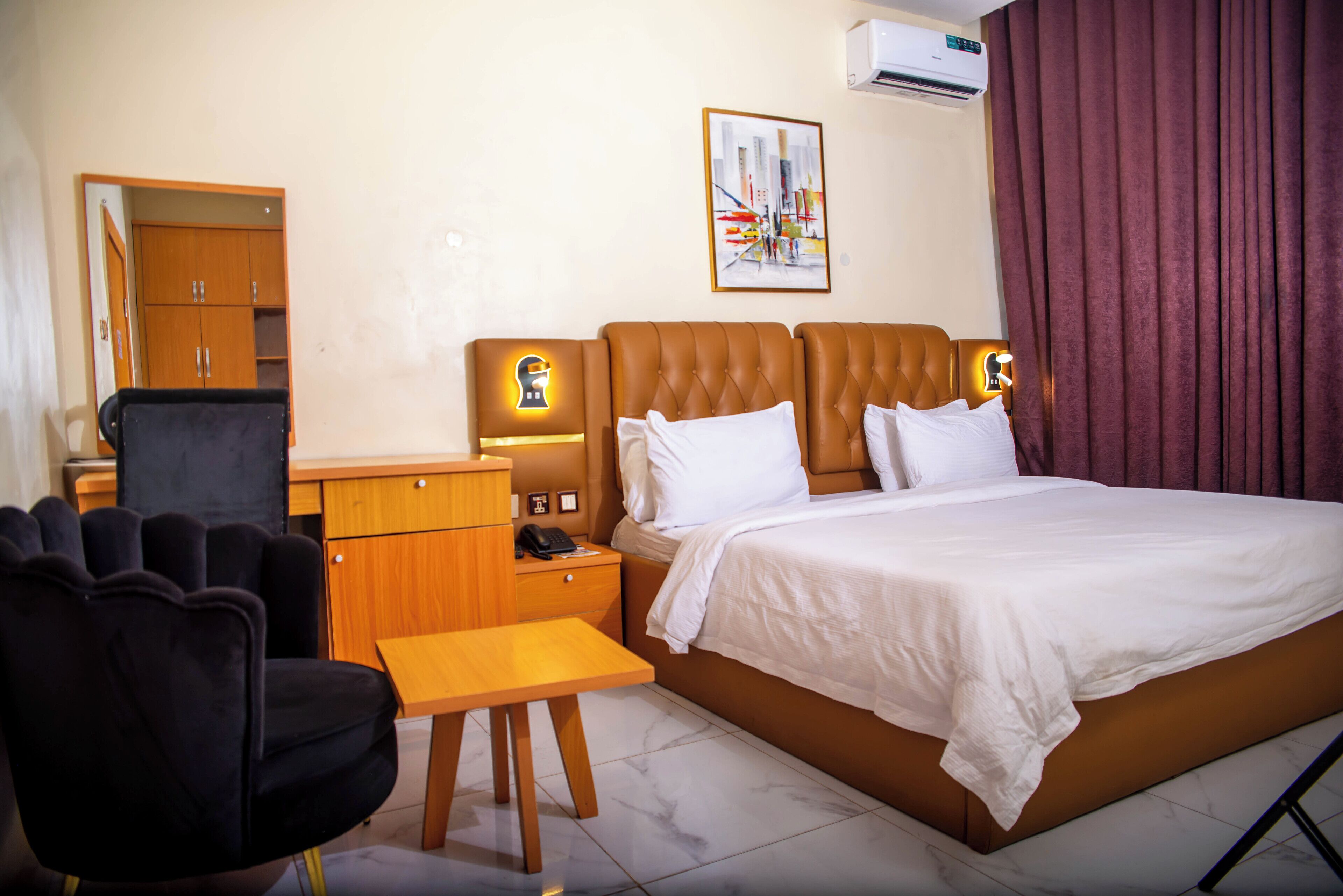 A hotel room with a bed, a desk, a chair, a wardrobe, and a painting on the wall.