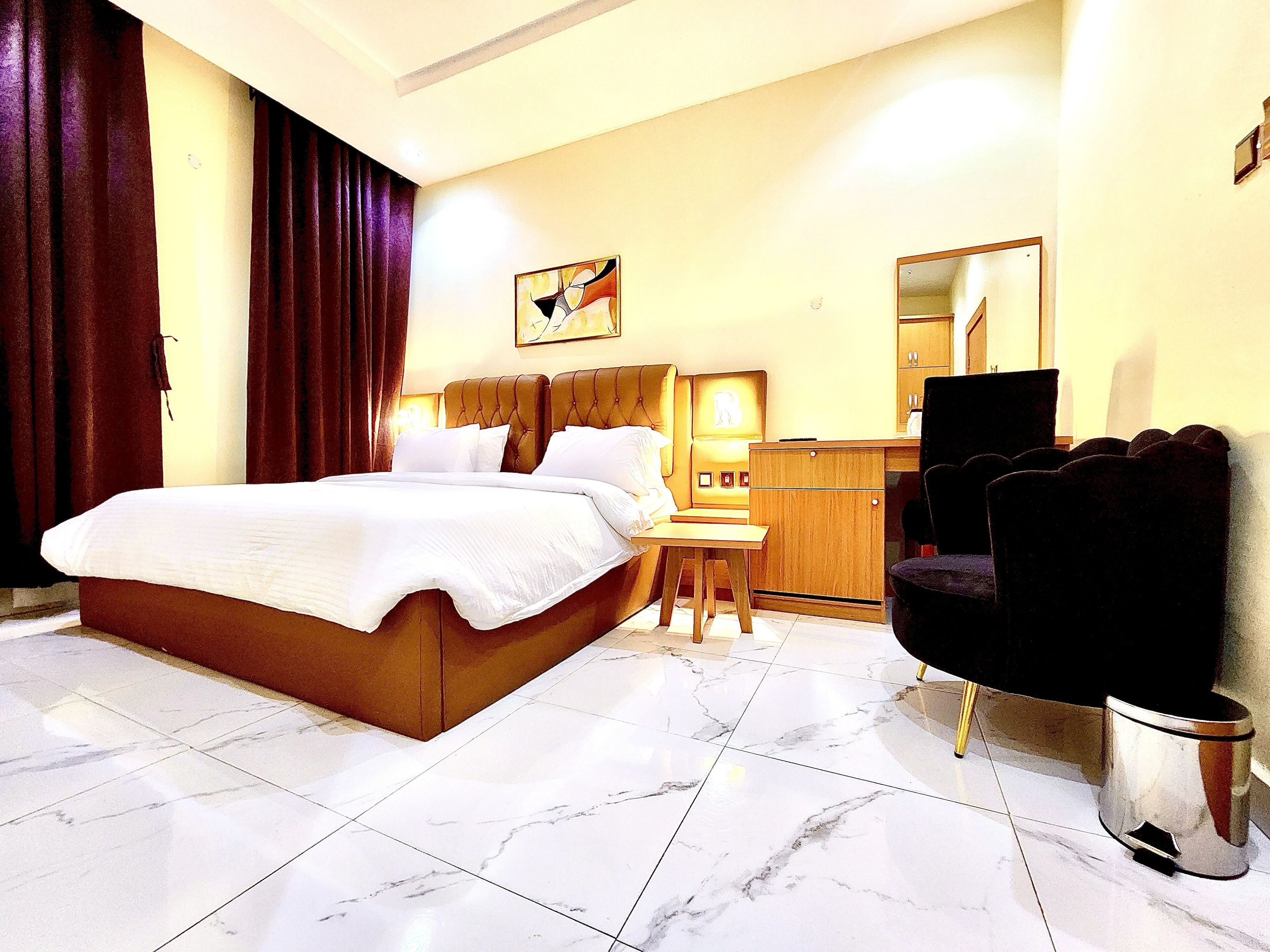 A hotel room with a large bed, a wooden desk, a chair, a trash bin, and a painting on the wall.