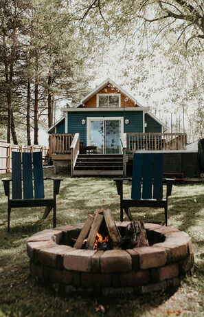 A cabin with a deck, two Adirondack chairs, and a fire pit.