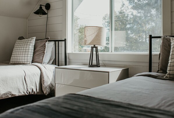A bedroom with two beds, a bedside table with a lamp, and a window with a view of trees.