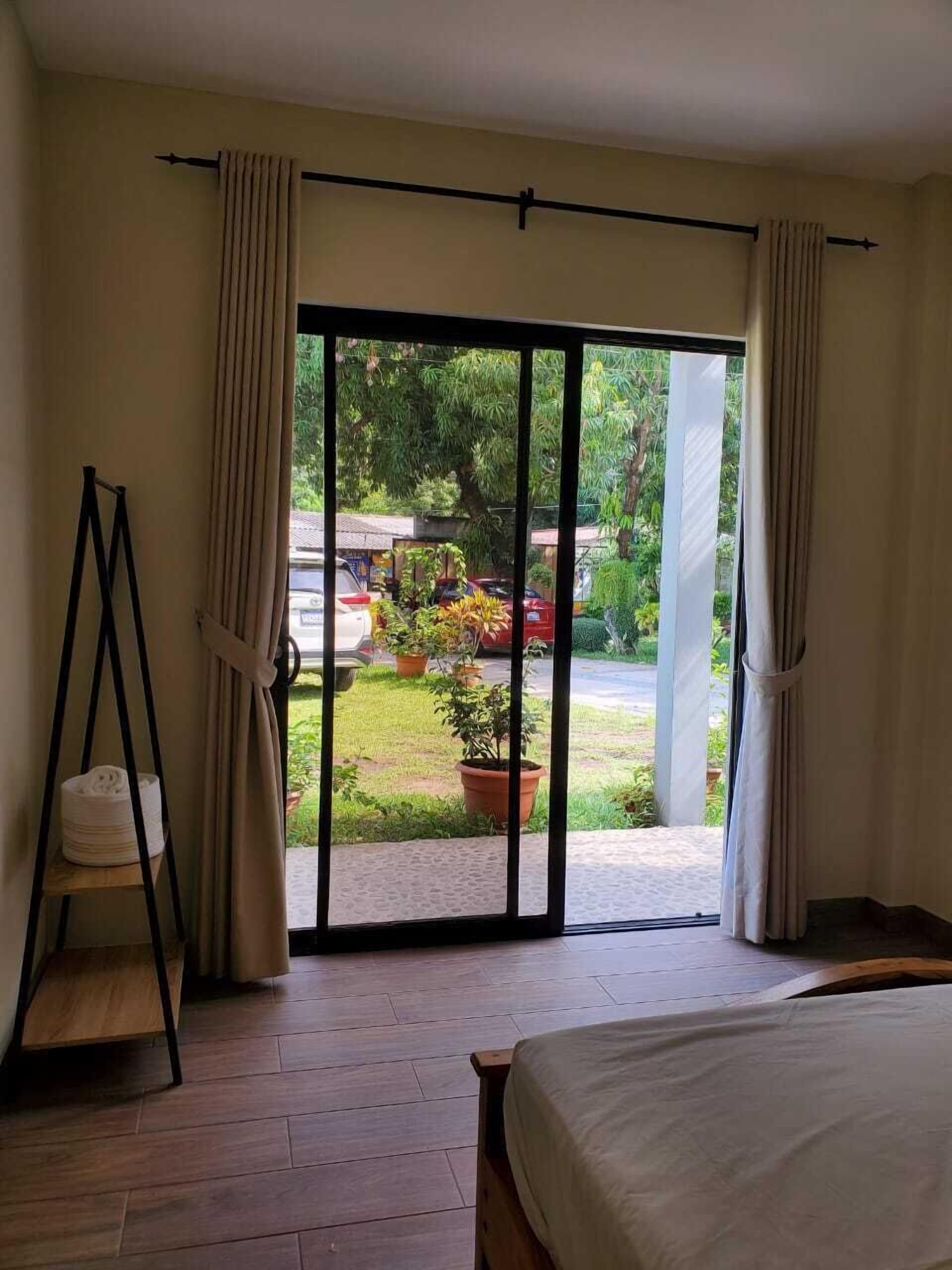 Un dormitorio con cama, piso de madera y vistas a un jardín con plantas en macetas y un coche estacionado a través de un ventanal corredero.