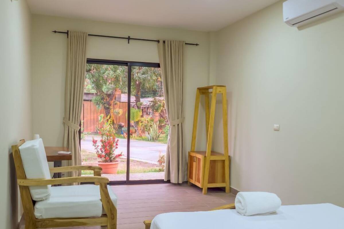 A hotel room with a bed, a wooden chair, a wooden table, a wooden ladder, and a view of the outdoors.