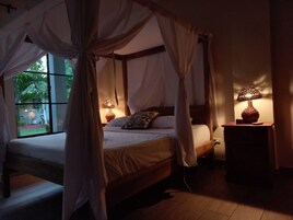 A four-poster bed with a canopy, two bedside lamps, and a view of greenery outside.