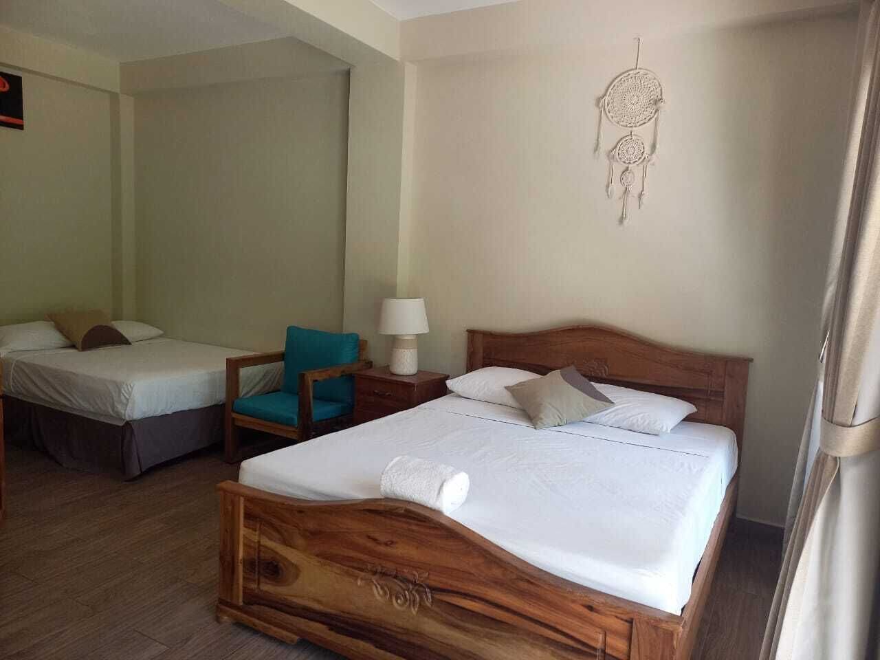 A hotel room with a large wooden bed, two smaller beds, a teal chair, a nightstand with a lamp, and a dreamcatcher on the wall.