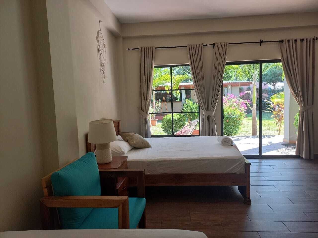 Une chambre à coucher avec un lit, une table de chevet en bois, une lampe et une vue sur un jardin grâce à de grandes fenêtres.