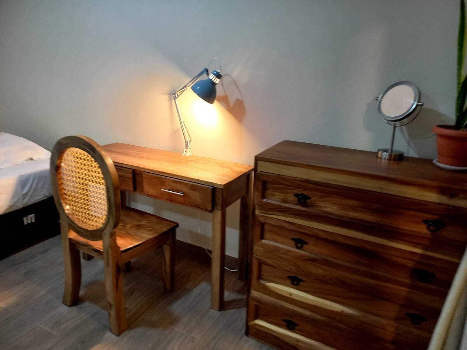 A wooden desk with drawers, a chair, a desk lamp, a small round mirror, and a potted plant.