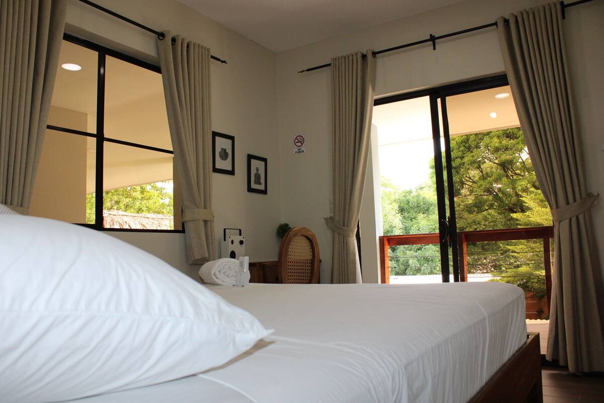 A bedroom with a large bed, two windows with curtains, a small table with a lamp, and a wall-mounted picture.