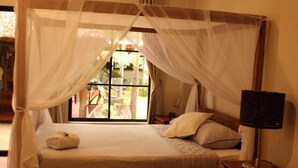 Un dormitorio con una cama bajo un dosel, una mesita de noche con una lámpara y vistas al exterior.