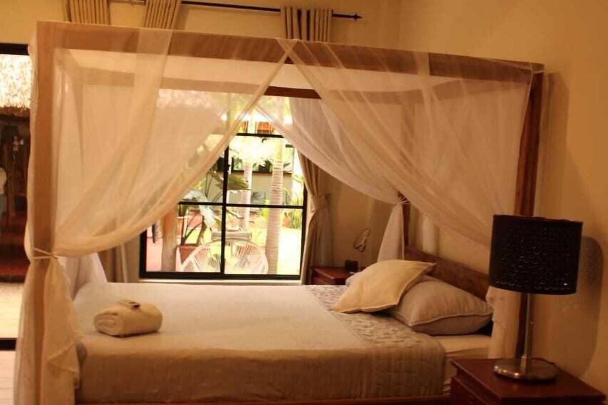 A bedroom with a bed under a canopy, a nightstand with a lamp, and a view of the outdoors.