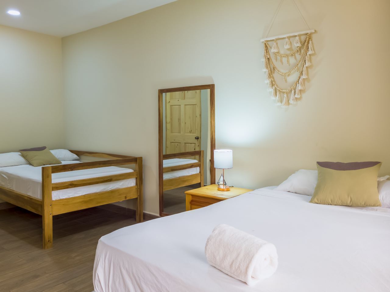Quarto com duas camas de madeira, uma mesa de cabeceira com luminária, um espelho e uma decoração de macramé pendente.