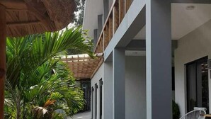 Un paseo techado con plantas en macetas, una silla blanca y un edificio con varios pisos y balcones.