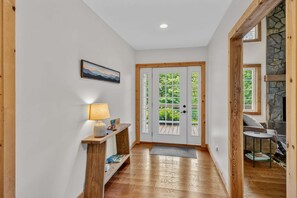 A hallway with a wooden floor, a white door with a view of greenery, a lamp on a wooden console, and a stone wall with a window.