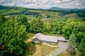 A rural house with a large porch, surrounded by dense trees and rolling hills in the background.