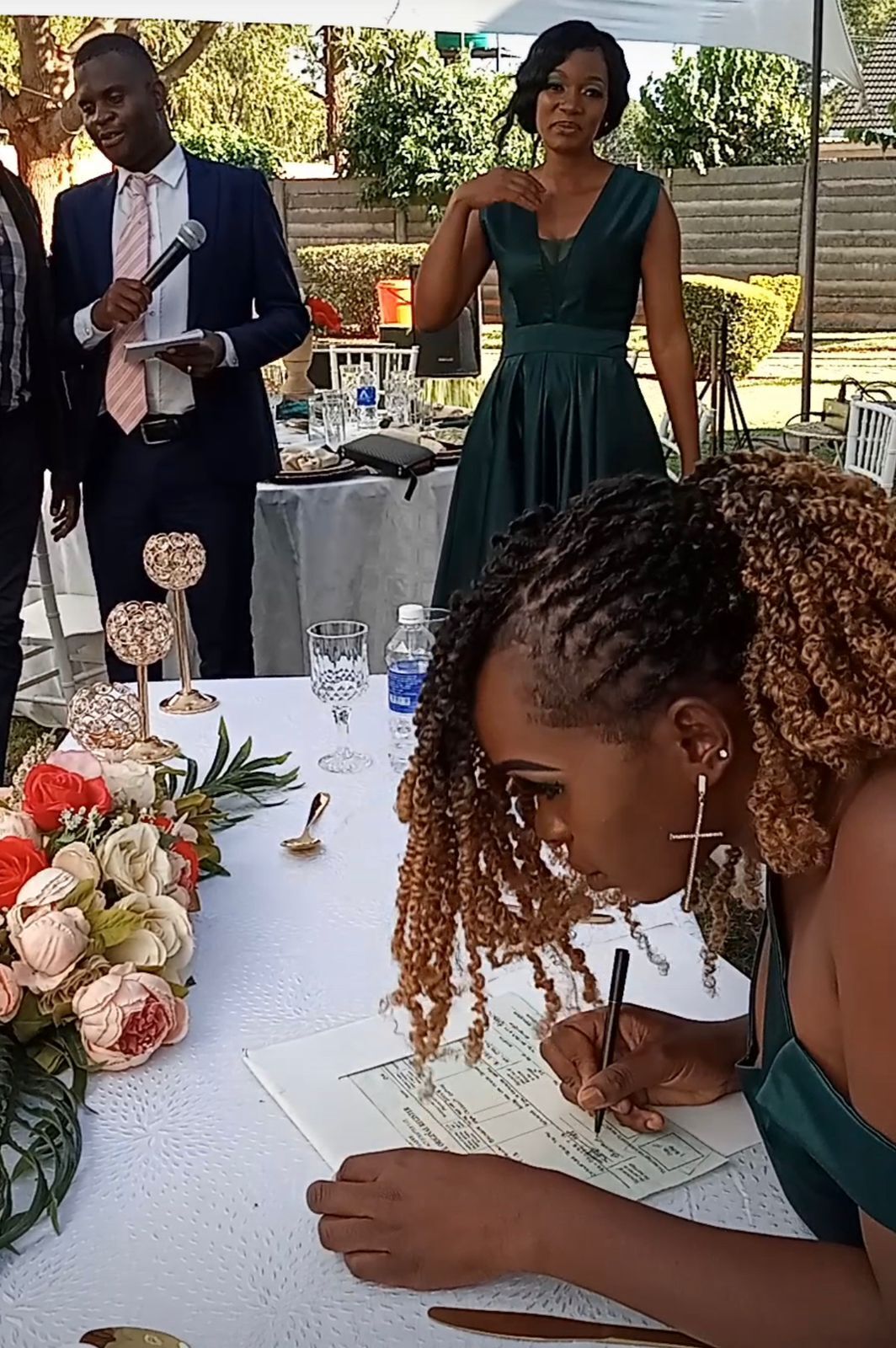 A person signing a document at a table with a floral arrangement and a microphone stand in the background.