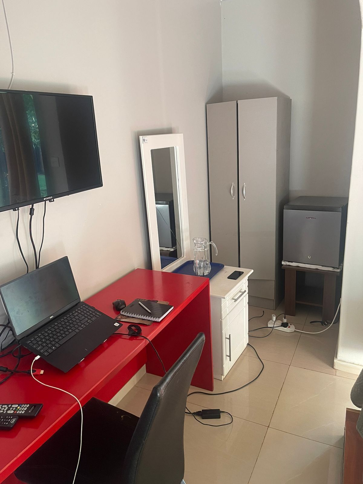 A small, well-lit room with a red desk, a laptop, a mirror, a wardrobe, and a small refrigerator.