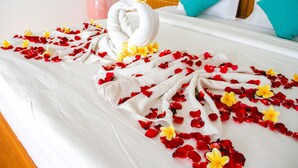 Un letto con lenzuolo bianco, petali di fiori rossi e gialli, e due asciugamani bianchi disposti a forma di cuore.